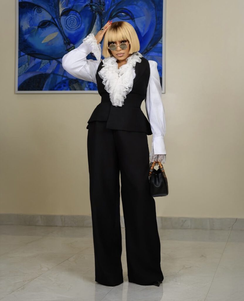 Rita Dominic in a black peplum waistcoat layered over a ruffled white shirt with wide-leg trousers and gold-framed sunglasses — modern Nigerian glamour styles