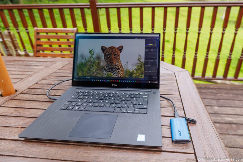 A laptop open on a wooden table outdoors, editing a leopard photograph in Lightroom, with a portable SSD plugged in alongside it for backup